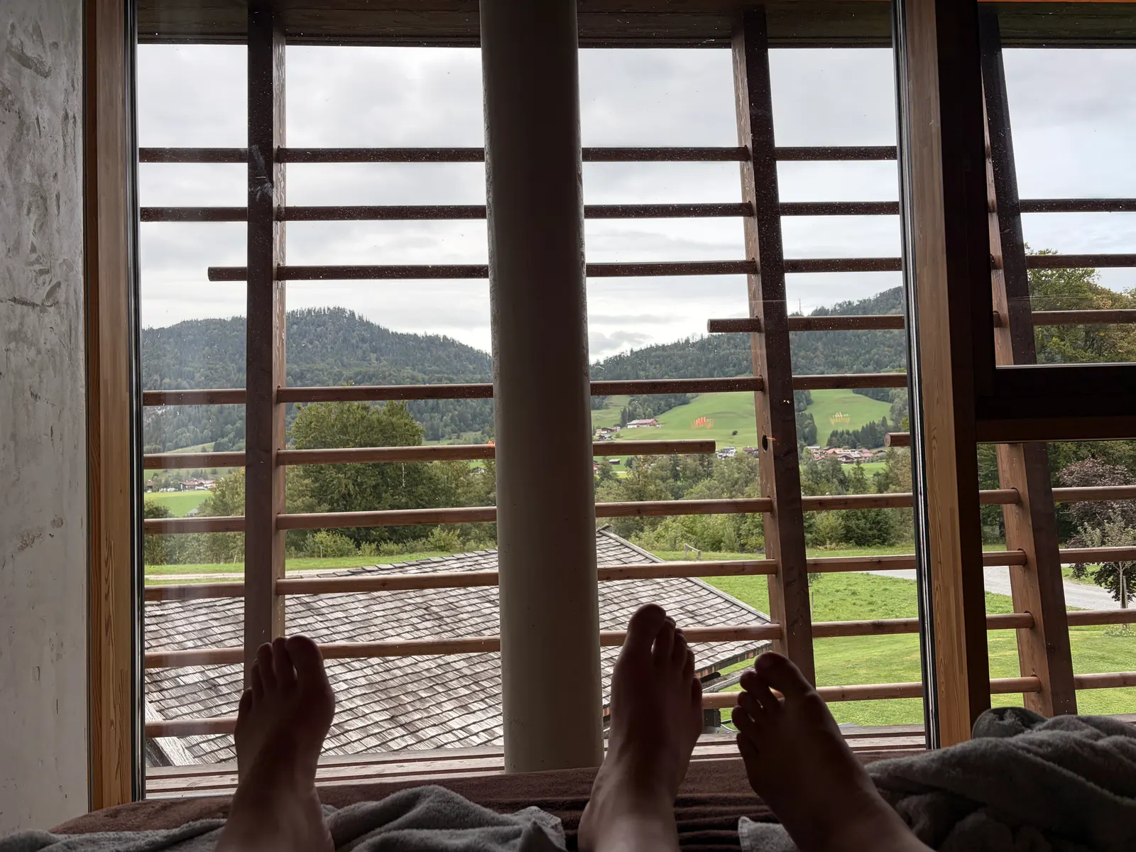 Blick vom Bett durch ein Fenster im Spa-Bereich vom Gut Steinbach mit sichtbaren Füßen auf Wiesen, Häuser und bewaldete Hügel bei bewölktem Himmel.