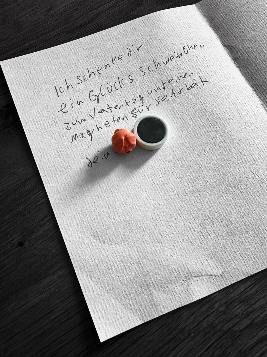 Handgeschriebene Vatertagskarte auf strukturiertem Papier mit einem kleinen rosa Glücksschweinchen und einem runden Magneten darauf. Die Nachricht lautet: „Ich schenke dir ein Glücksschweinchen zum Vatertag und einen Magneten für die Arbeit. “