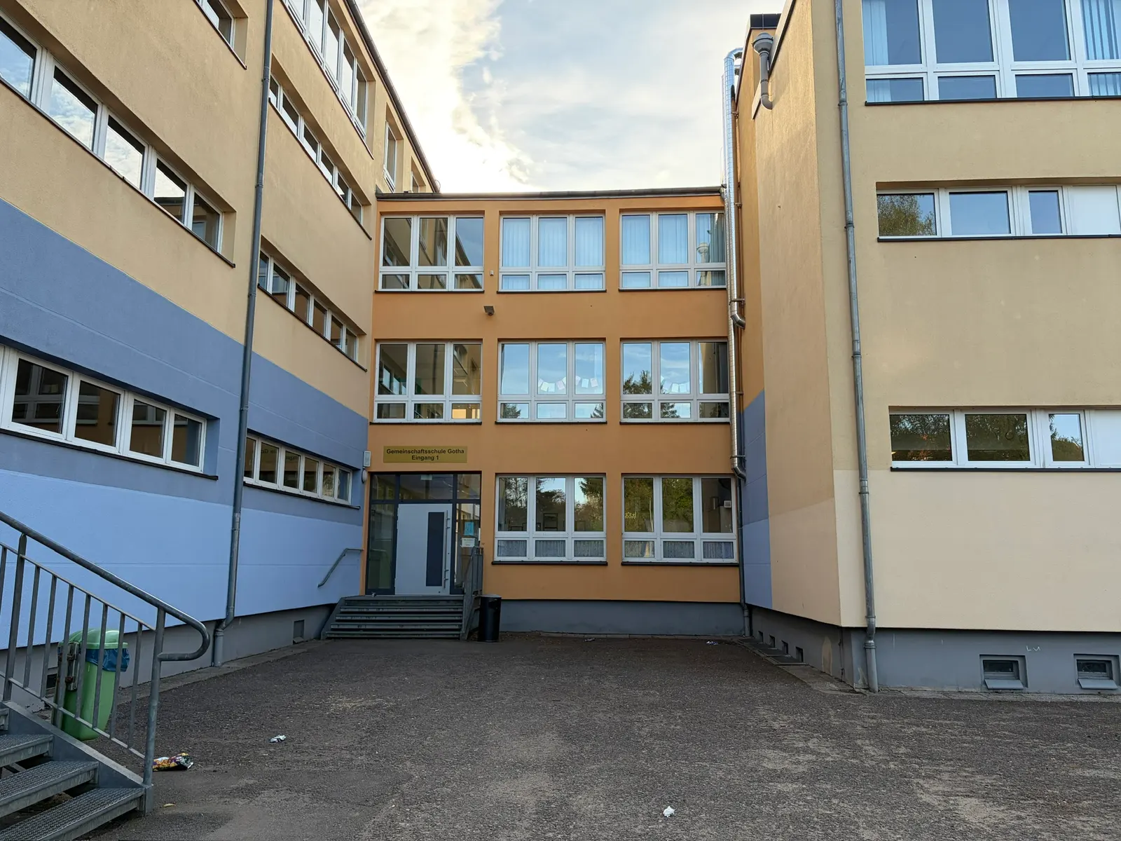 Innenhof einer Schule mit beigefarbenen Gebäudefassaden, vielen Fenstern, Eingangstreppe und leerer Asphaltfläche.