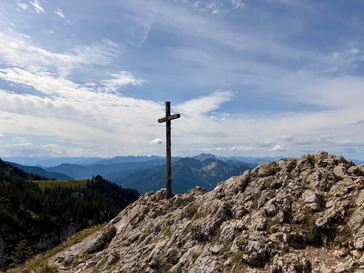 Gipfelkreuz auf felsigem Berggrat vor weitem Alpenpanorama unter blauem, leicht bewölktem Himmel
