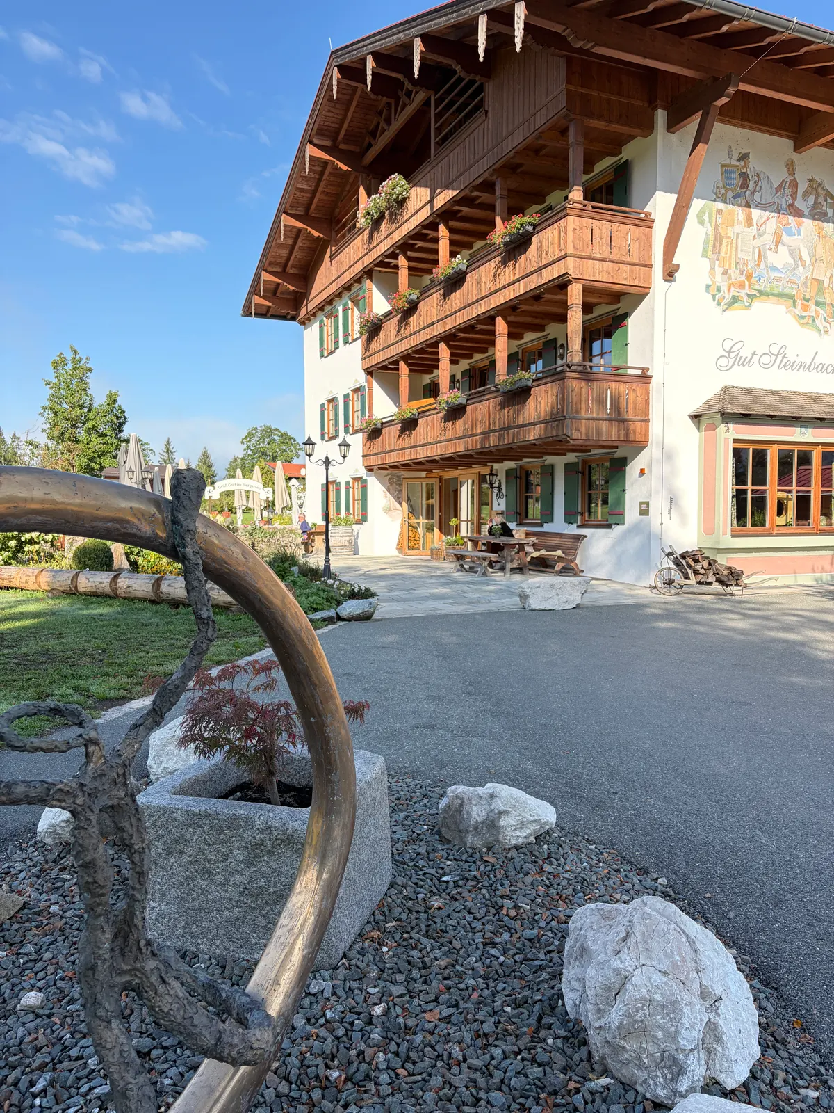 Gut Steinbach im alpinen Stil mit Holzbalkonen und Sonnenterrasse, davor Kiesfläche, Felsen und eine gebogene Holzskulptur bei blauem Himmel.
