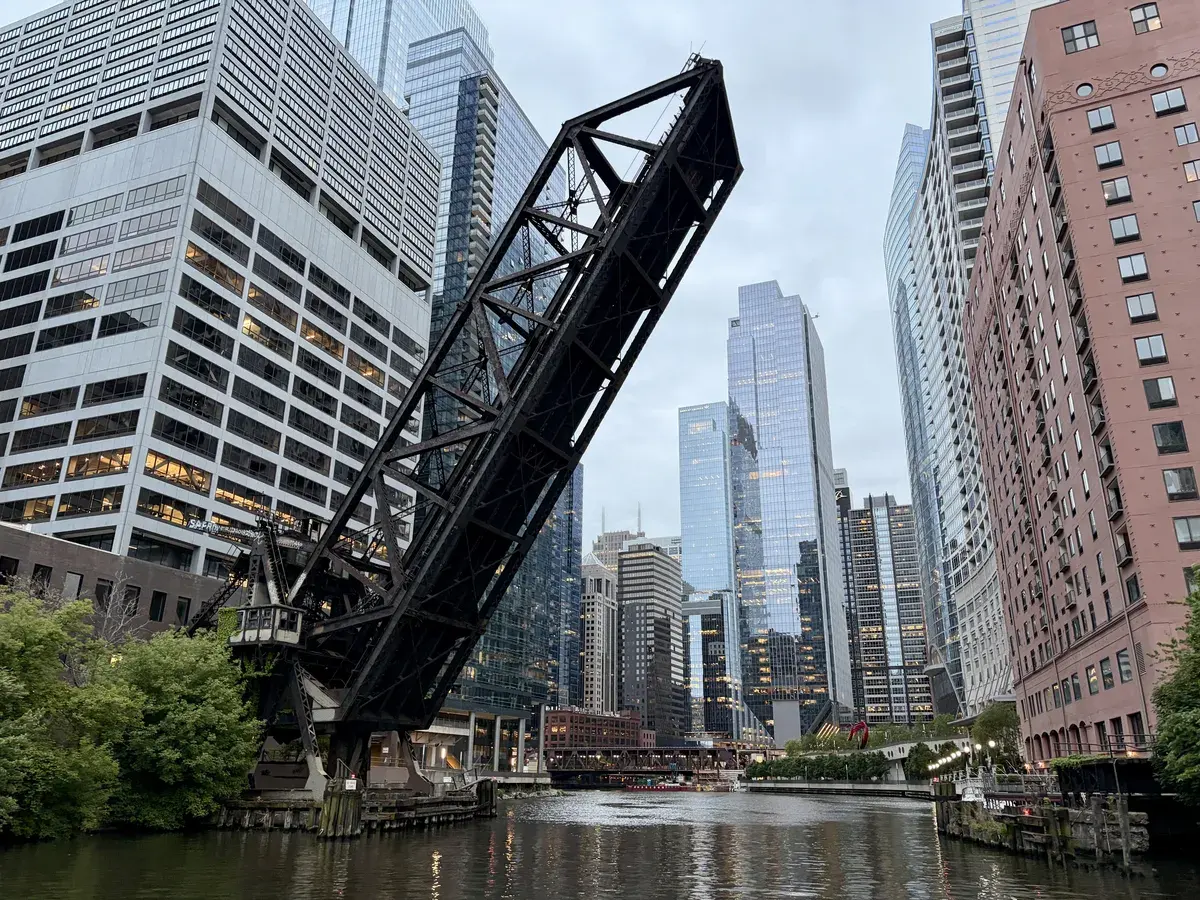 Eine große, schwarze Drehbrücke ist in einer senkrechten Position über einem Stadtwasserweg angehoben. Umgeben von hohen modernen Wolkenkratzern spiegelt sich die Architektur im glatten Wasser wider.