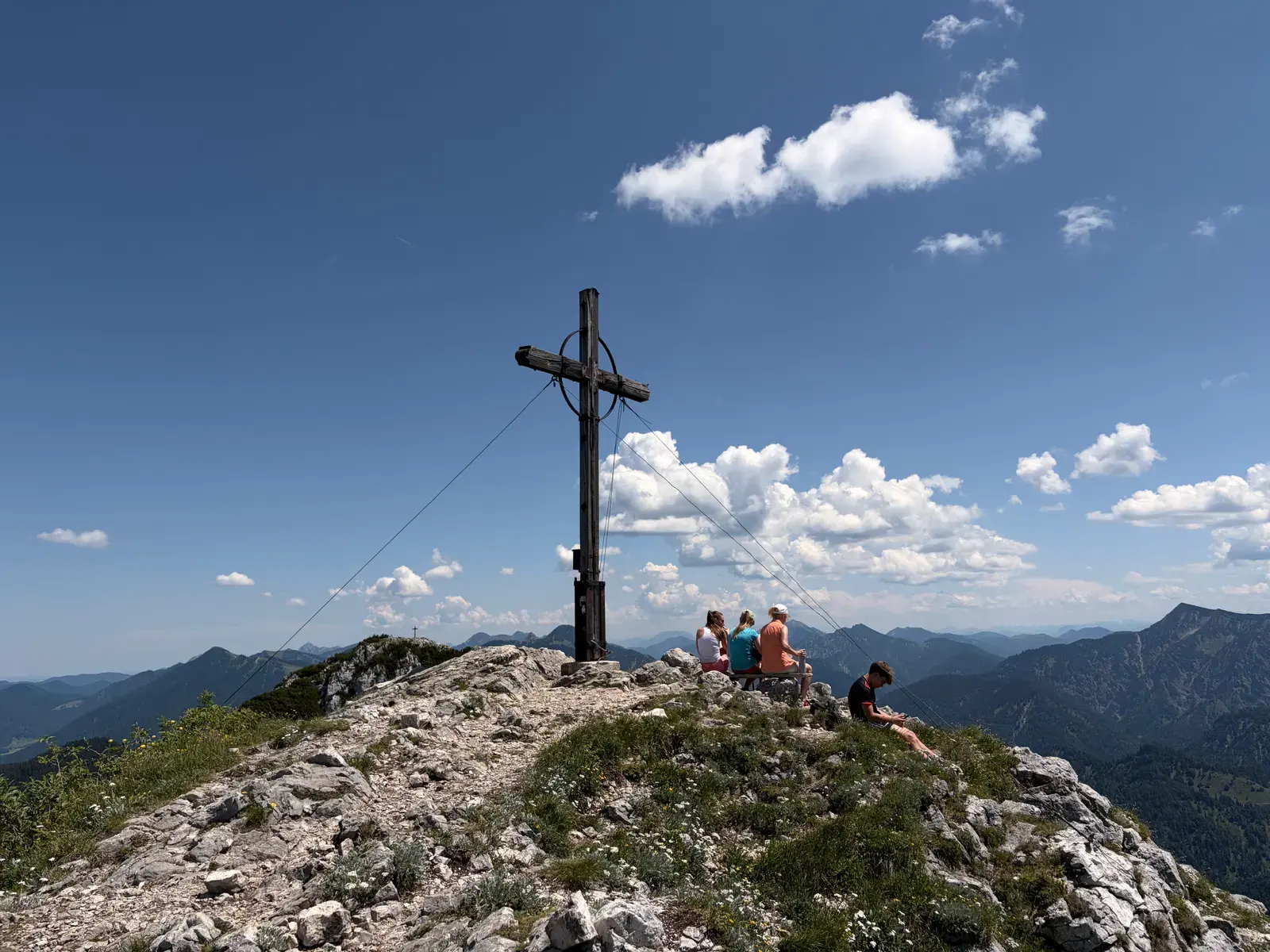 Gipfelkreuz des Roßstein auf einem felsigen Bergkamm , daneben sitzen mehrere Wanderer. Im Hintergrund erstreckt sich bei klarem Himmel die Berglandschaft rund um die Tegernseer Hütte.