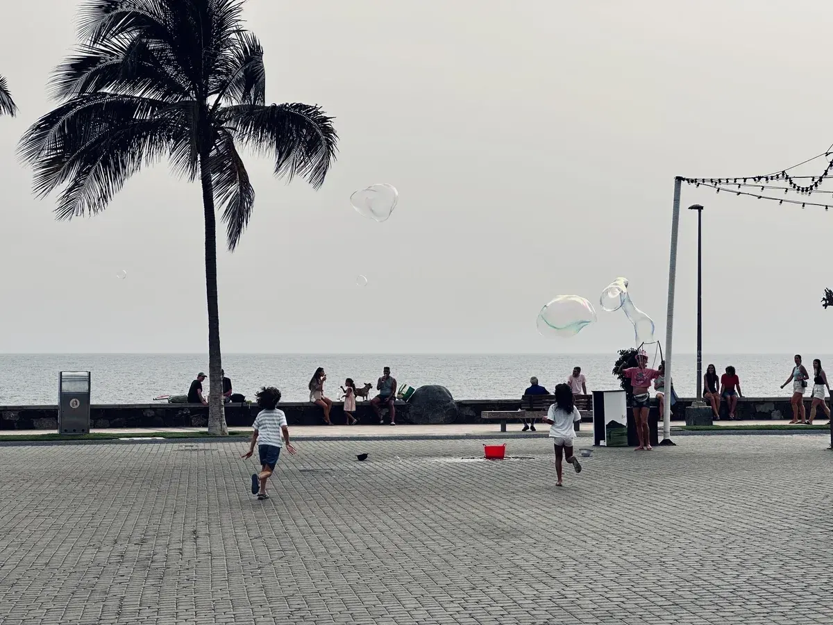Kinder spielen mit Seifenblasen an der Strandpromenade von Maspalomas, während Leute auf einer Mauer sitzen, und Palmen stehen im Hintergrund.