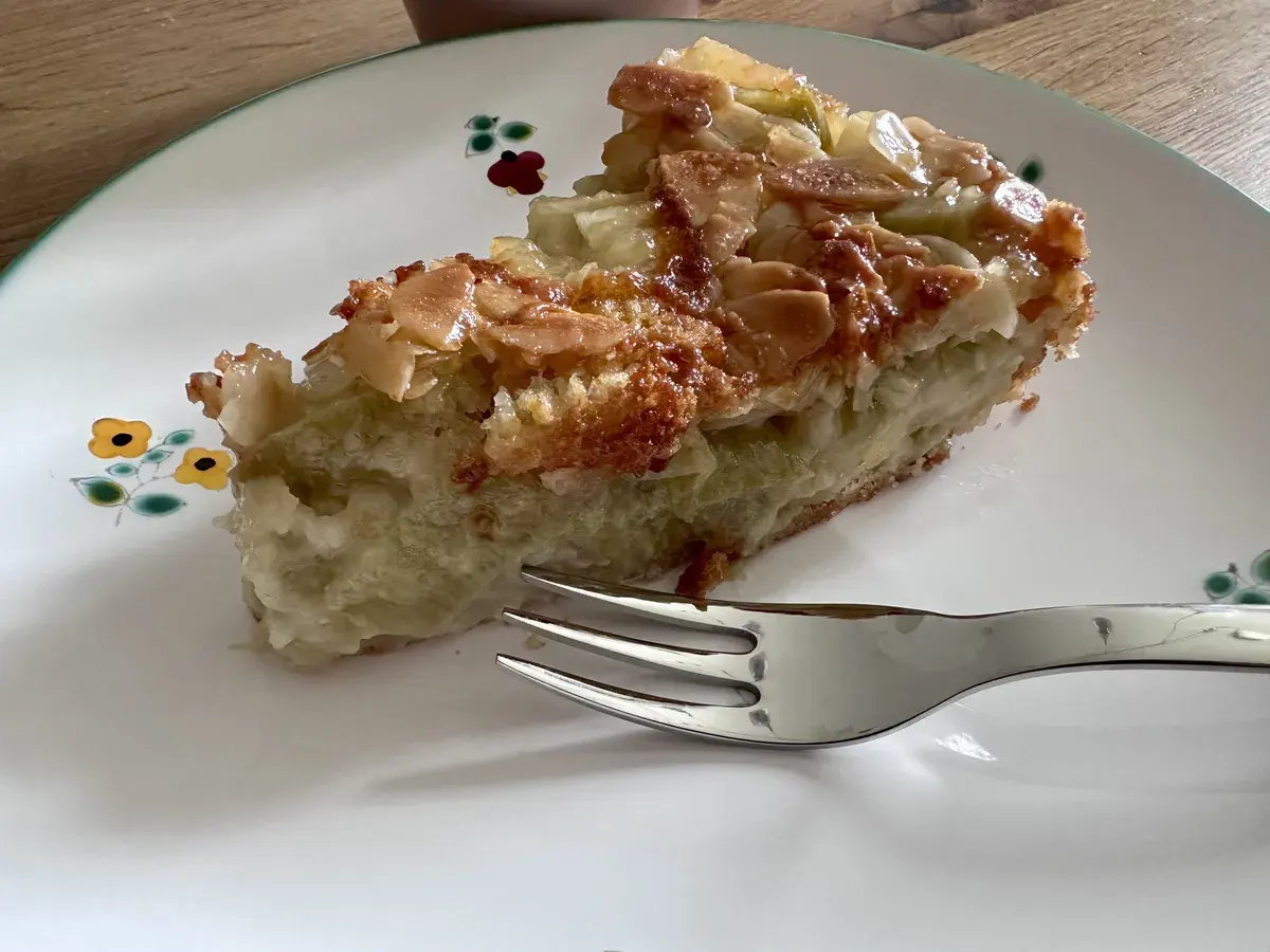 Ein Stück Kuchen mit einer goldbraunen, knusprigen Oberseite, das auf einem weißen Teller mit bunten Blütenmustern angerichtet ist. Eine Gabel liegt daneben, während im Hintergrund ein Becher zu sehen ist.