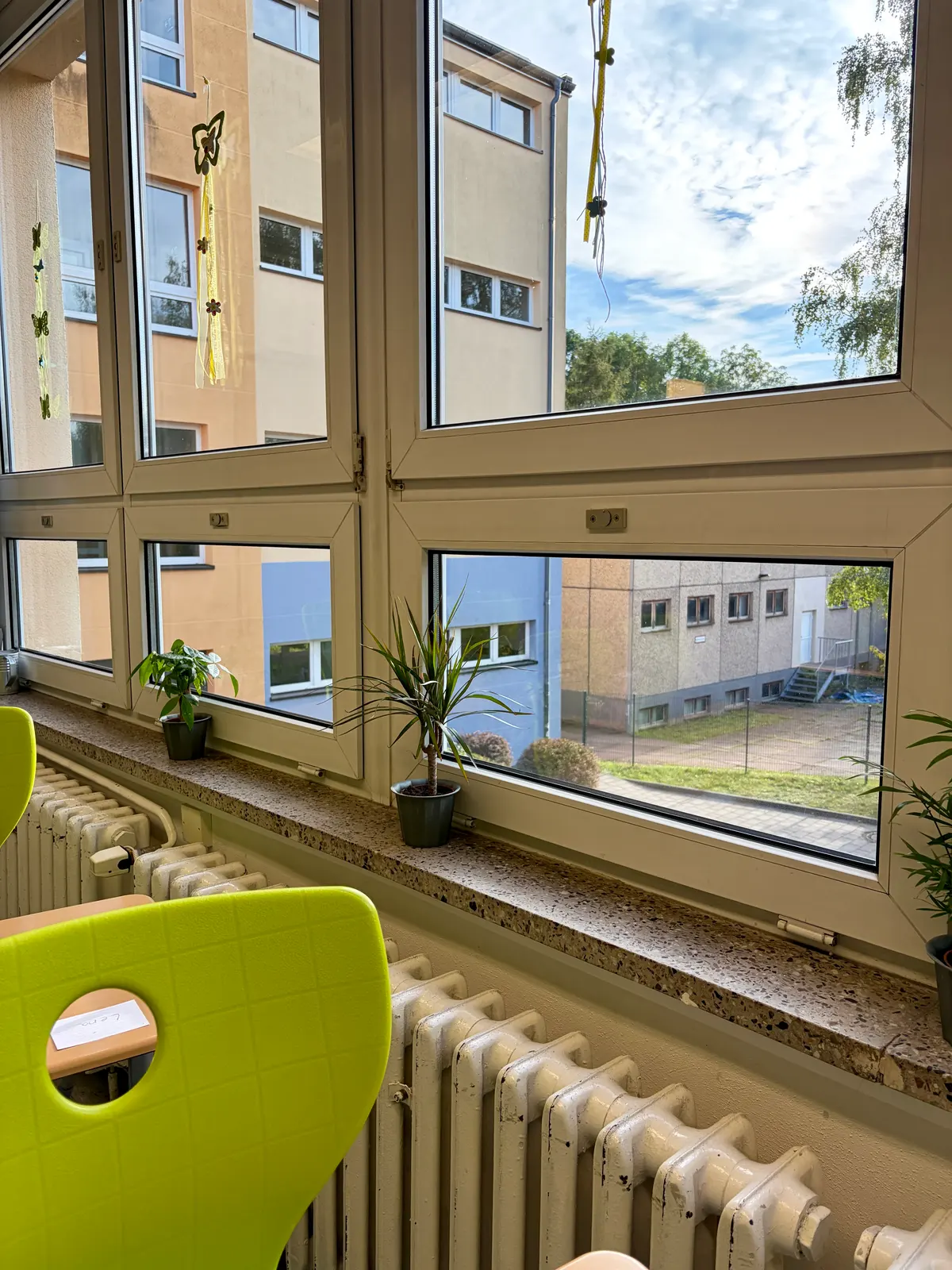 Geöffnetes Klassenzimmerfenster mit Blick auf Schulgebäude, Fensterbank mit Pflanzen und Heizkörper davor