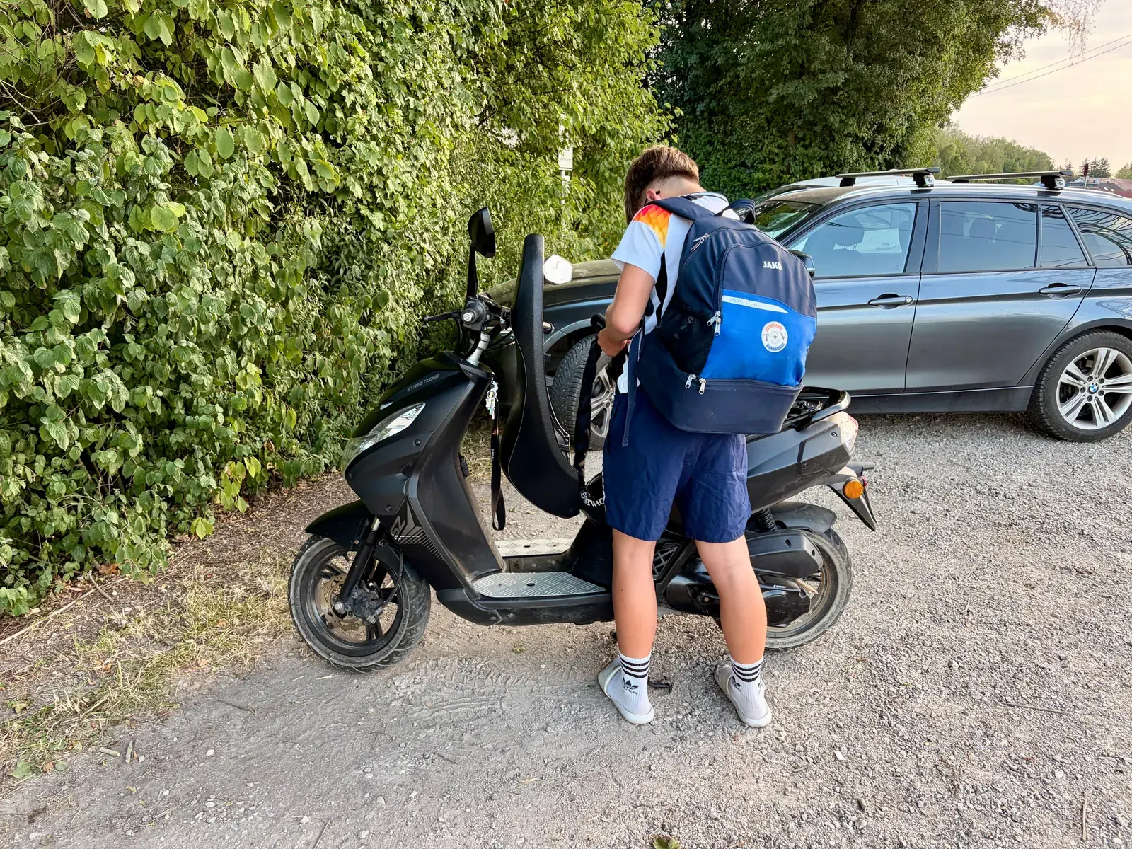 Ein Jugendlicher mit Rucksack steht neben einem schwarzen Roller auf einem Kiesweg und hält einen Helm in der Hand. Im Hintergrund stehen ein Auto und dichtes grünes Gebüsch.