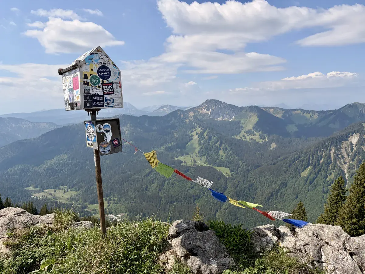 Ein kleiner Blechkasten mit Aufklebern steht auf einem felsigen Gipfel und verwahrt das Gipfelbuch, umgeben von einer beeindruckenden Berglandschaft. Bunte Gebetsfahnen wehen im Wind und verleihen der Szene einen spirituellen Charakter.