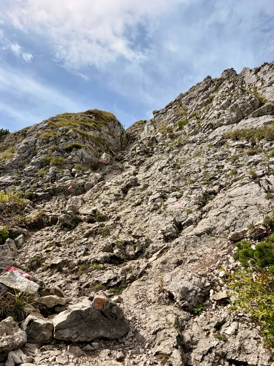 Steiler felsiger Bergpfad mit roten Markierungen zwischen schroffen Kalkfelsen und Graspolstern unter blau bewölktem Himmel