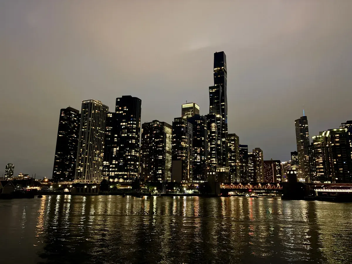 Eine Stadtansicht mit modernen Hochhäusern, die bei Nacht beleuchtet sind. Das Wasser im Vordergrund spiegelt die Lichter der Gebäude wider, während der Himmel eine graue Färbung hat.