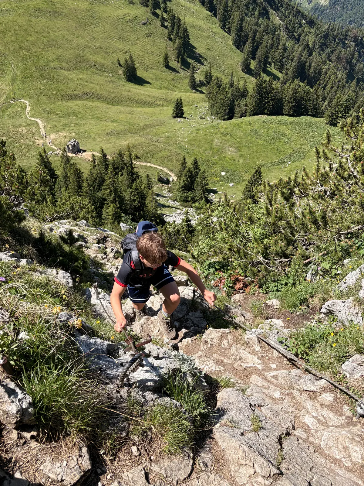 Ein Wanderer kraxelt in steilem, felsigem Gelände zur Tegernseer Hütte hinauf. Unterhalb liegen grüne Almwiesen, Wald und ein schmaler Bergpfad.