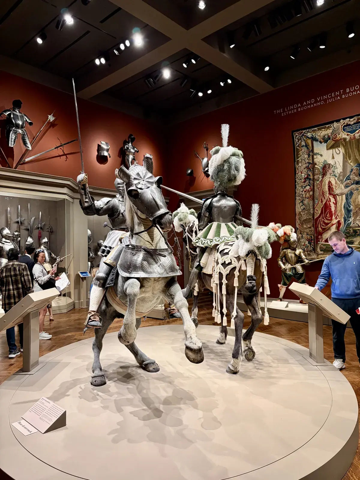 Zwei mit Rüstungen ausgestattete Ritter auf Pferden stehen in einem Museum. Der Hintergrund zeigt eine Wand mit weiteren ritterlichen Rüstungen und historischen Waffen, während Besucher das Exponat betrachten.