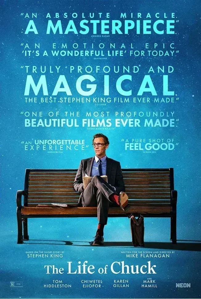Filmplakat zu The Life of Chuck: Ein Mann im Anzug sitzt auf einer Parkbank vor blauem Hintergrund, darüber stehen große Pressestimmen wie „A Masterpiece“ und „Magical“.