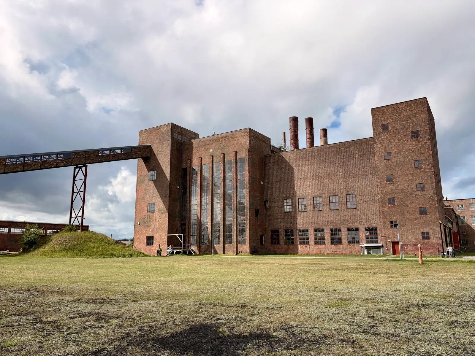 Backsteinbau des ehemaligen Kohlekraftwerks Peenemünde mit Schornsteinen, Förderbrücke und Wiese im Vordergrund unter wolkigem Himmel