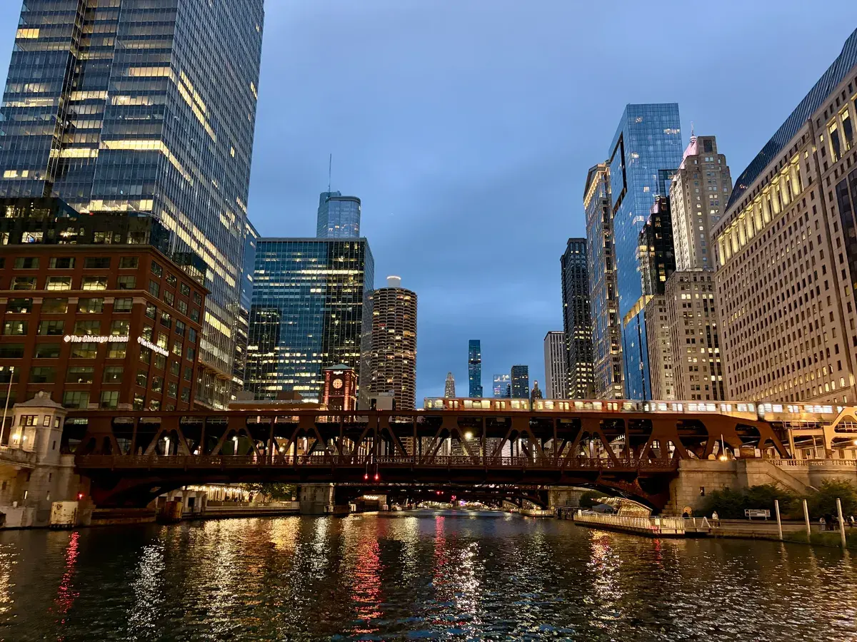 Eine elegante Brücke über einem ruhigen Fluss umgeben von hohen modernen Gebäuden und Wolkenkratzern in der Dämmerung. Das Wasser reflektiert die Lichter der Stadt und schafft eine stimmungsvolle Atmosphäre.