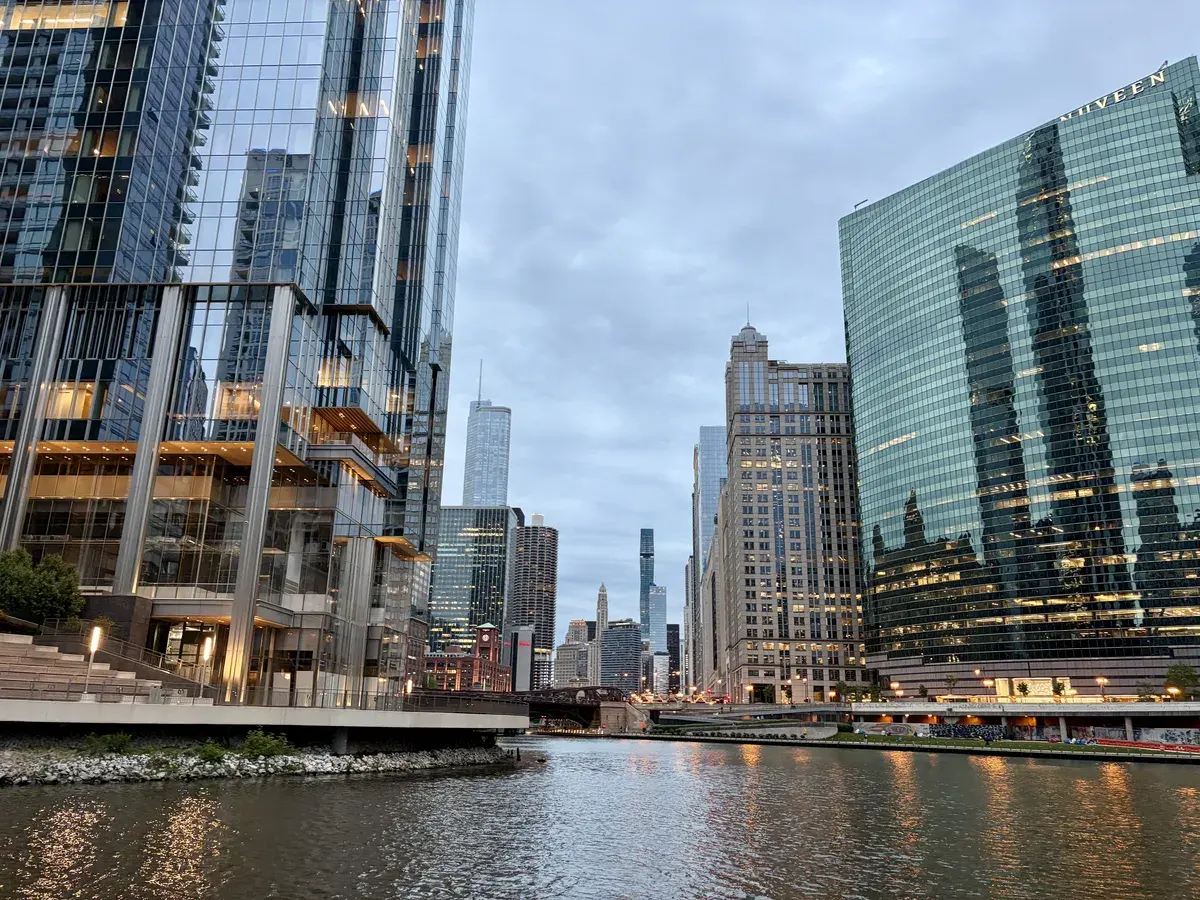Eine Sicht auf eine Stadtlandschaft mit modernen Wolkenkratzern, die sich im Wasser eines Flusses spiegeln. Der Himmel ist bewölkt, und es sind bereits Lichter an den Gebäuden eingeschaltet.