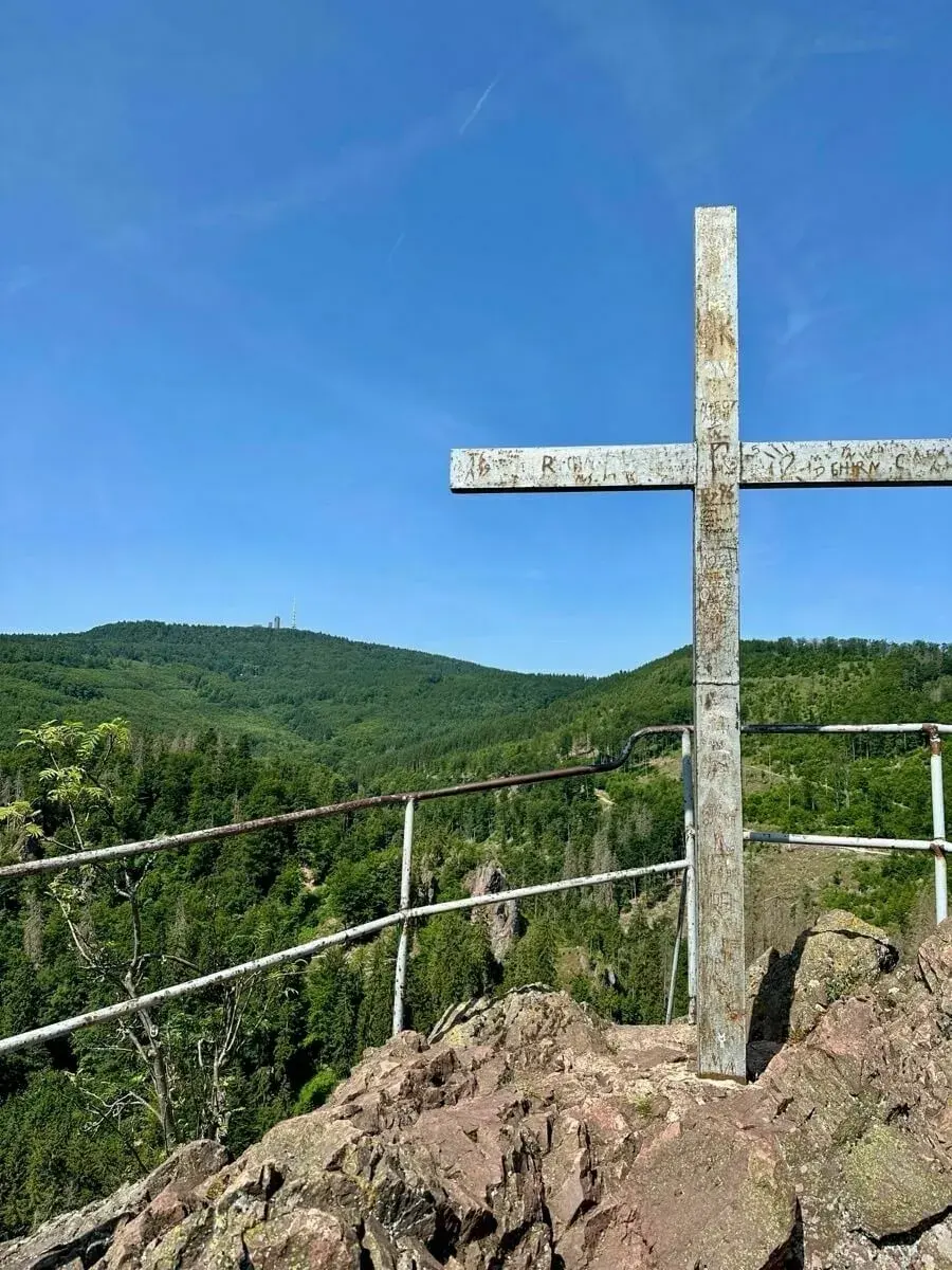 Ein großes Metallkreuz steht auf dem Aschenbergstein mit Aussicht auf den Inselsberg, bewaldete Hügel und blauen Himmel.