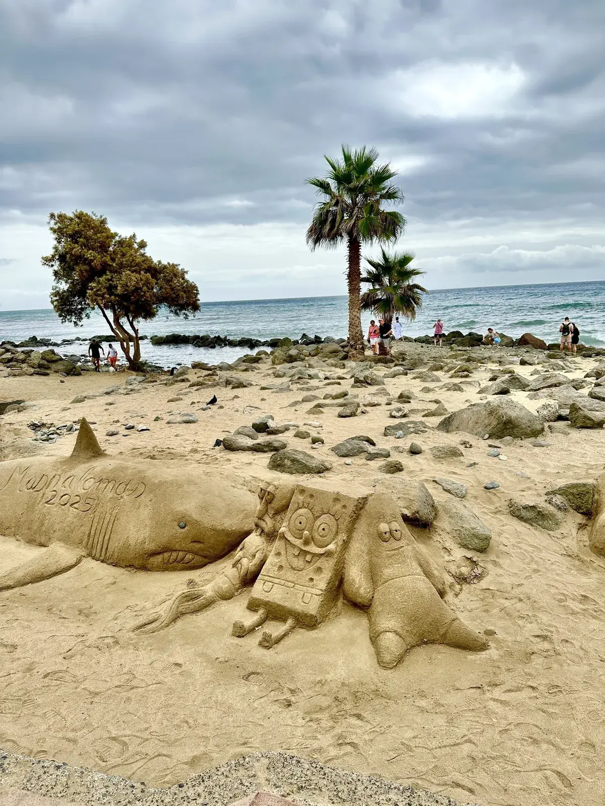 Ein Sandkunstwerk zeigt einen Hai, SpongeBob Schwammkopf und Patrick Star auf dem Strand in Maspalomas mit Palmen und bewölktem Himmel im Hintergrund.
