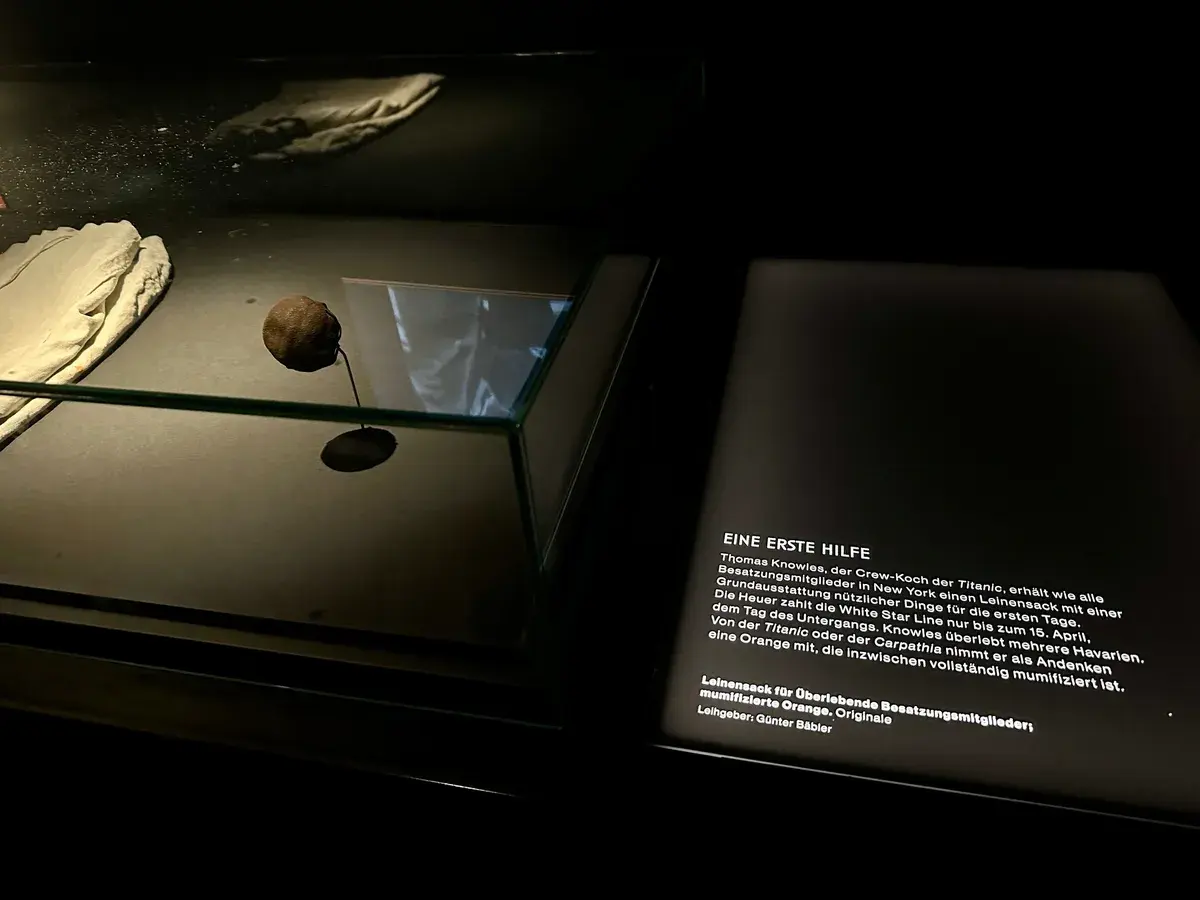 Eine Museumspräsentation zeigt eine mumifizierte Orange und ein Dokument mit Text über die Krise der Titanic, beschrieben in Deutsch.