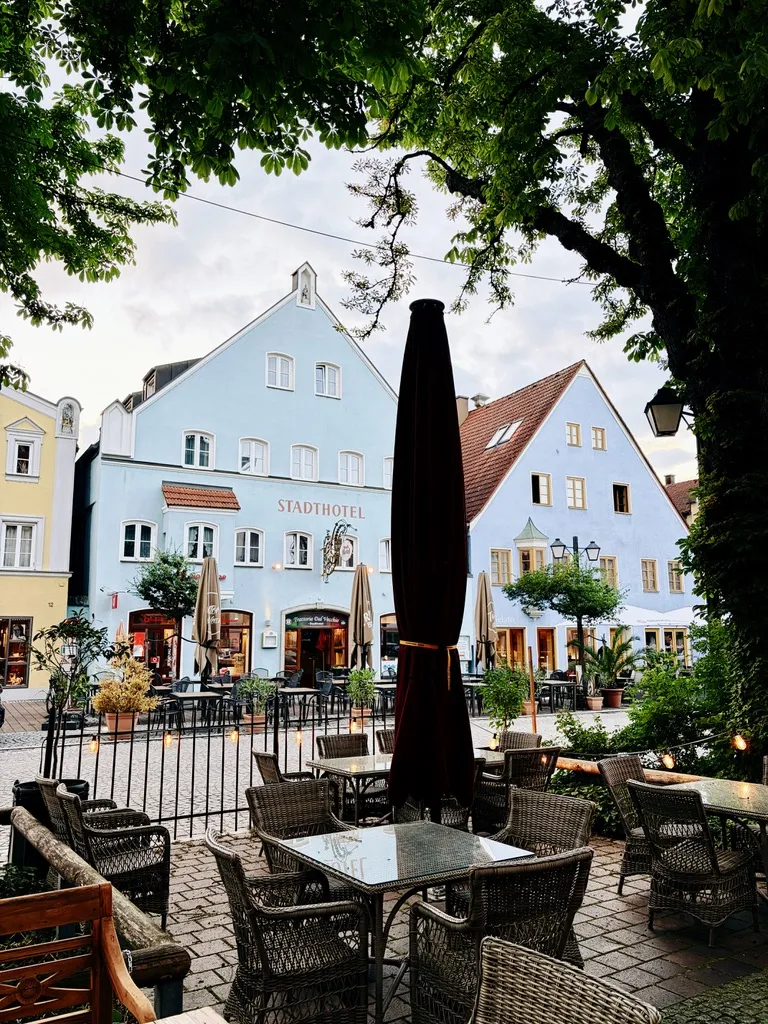 Caféterrasse mit Korbstühlen und geschlossenem Sonnenschirm unter Bäumen, dahinter pastellfarbene Giebelhäuser am Stadtplatz von Erding. Der Himmel ist bewölkt, die Szene wirkt wie eine ruhige Abendstimmung nach dem Regen.