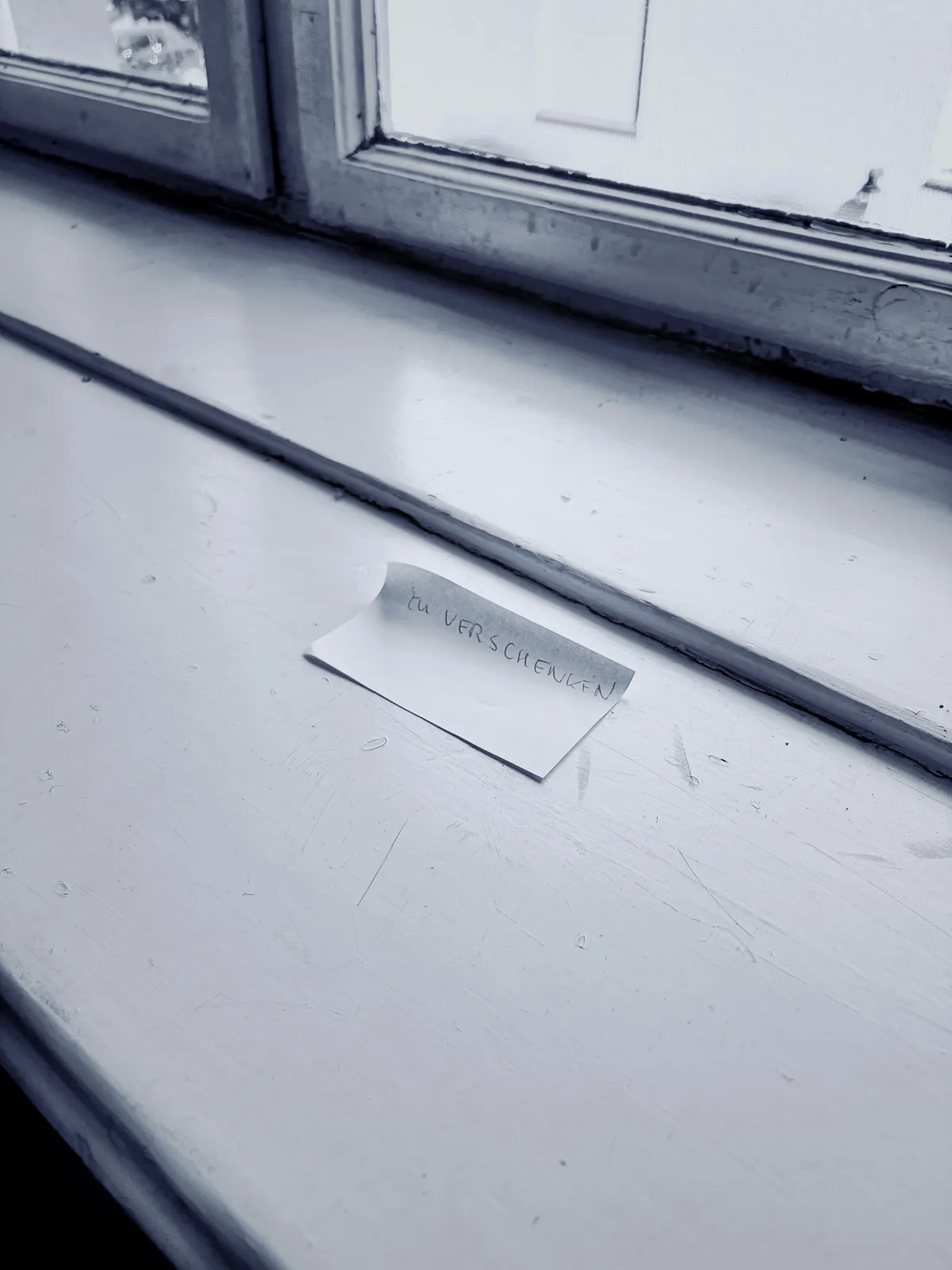Ein Stück Papier mit der handgeschriebenen Notiz „DU VERSCHIENEN“ liegt auf einer weißen Fensterbank, neben einem Fenster mit leicht verschwommenem Licht.