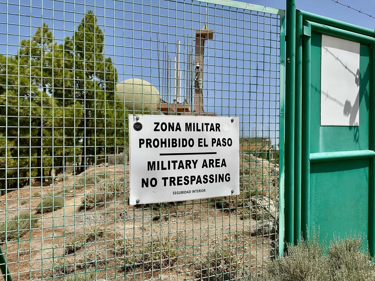Ein Schild mit der Aufschrift Military Area - No Trespassing hängt an einem Metallzaun, der ein Gelände mit militärischen Einrichtungen auf dem Pico de las Nieves umgibt.
