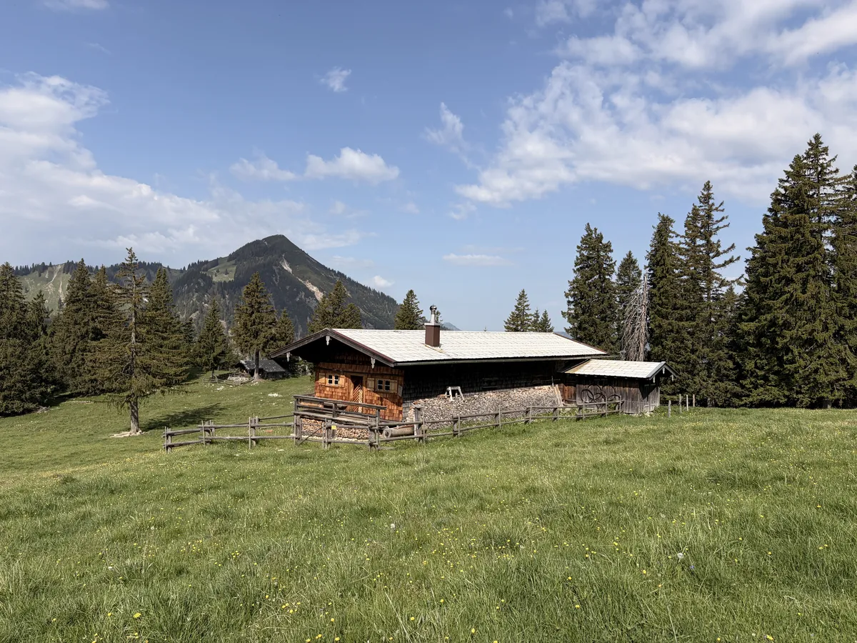 Ein rustikales Holzhaus steht auf einer Wiese, umgeben von grünen Bäumen und einem malerischen Berg im Hintergrund. Der Himmel ist teilweise bewölkt, mit einigen blauen Abschnitten.