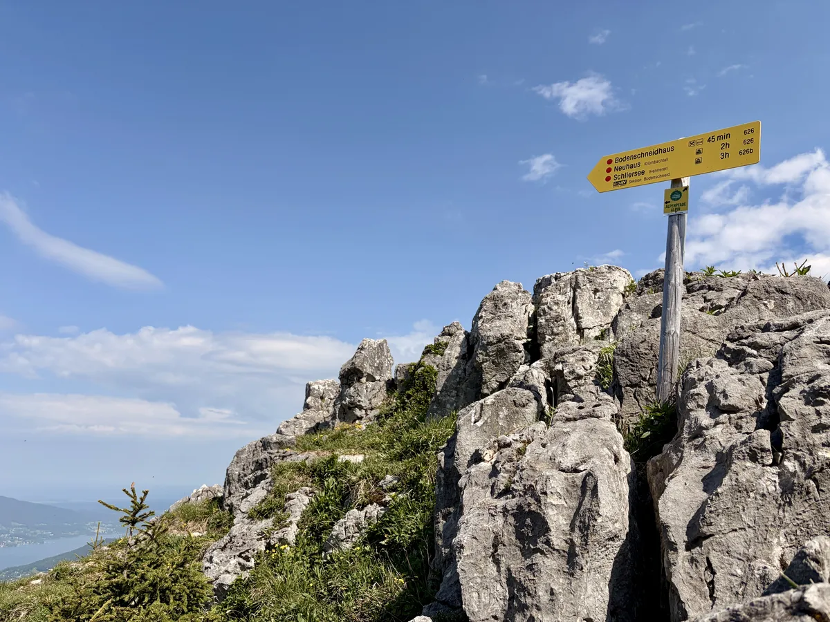 Ein gelbes Wegweiser-Schild steht an einer Felsenlandschaft auf einem Berg, mit blauen Himmel und wenigen Wolken im Hintergrund. Das Schild zeigt Entfernungen zu verschiedenen Wanderzielen an.