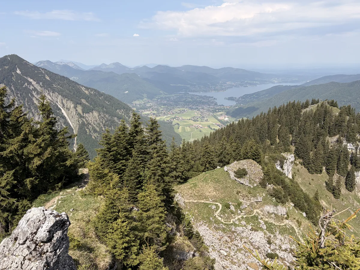 A weite Aussicht auf eine bergige Landschaft mit dichten Wäldern und dem ruhigen Tegernsee in der Ferne. Im Vordergrund sind schroffe Felsen und Wanderwege sichtbar, während die Berge und Täler bis zum Horizont reichen.