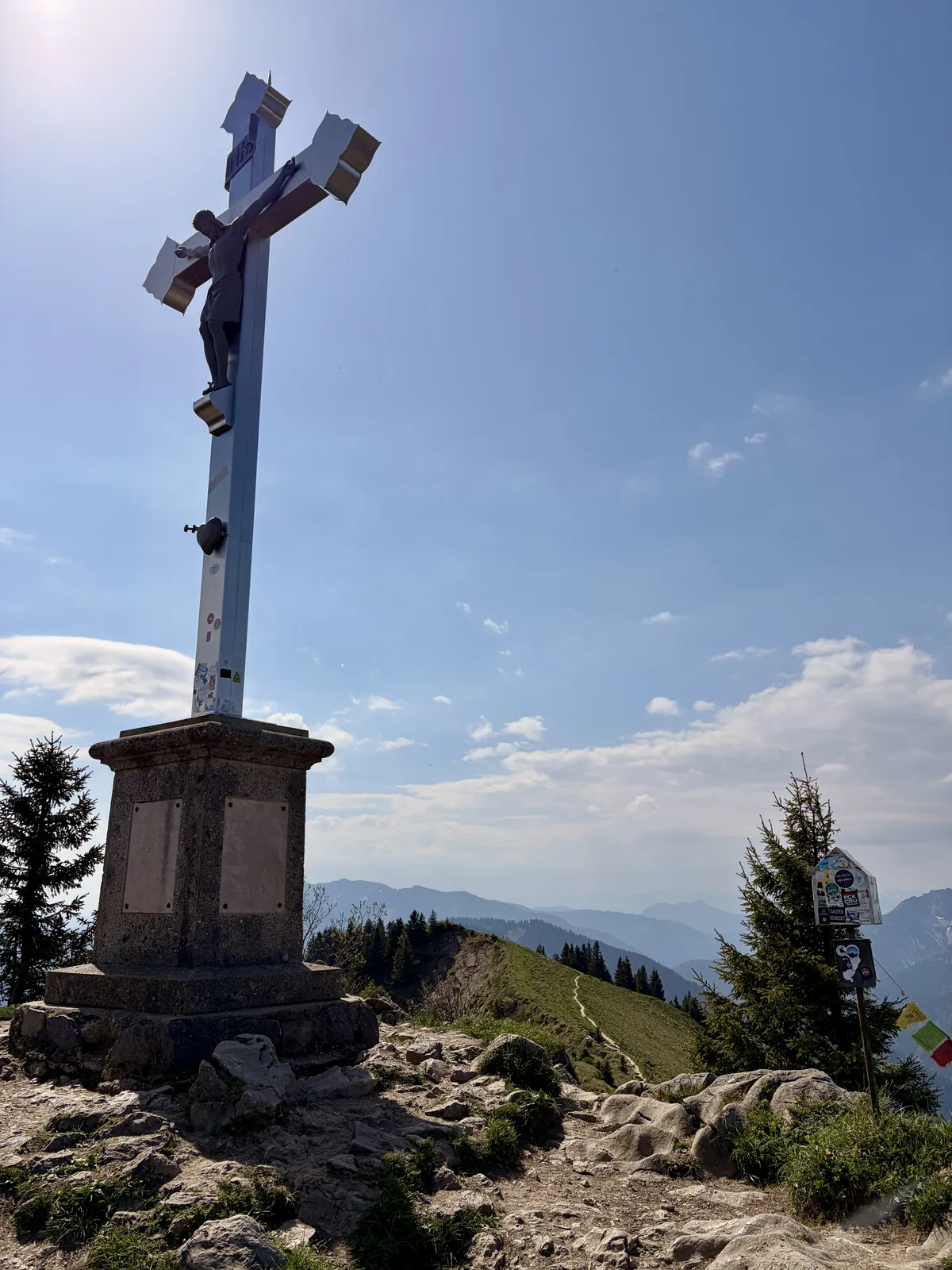 Ein hoher, silberner Kreuz auf einem steinernen Sockel steht auf einem Berggipfel, umgeben von Nadelbäumen. Im Hintergrund erstreckt sich eine hügelige Landschaft unter einem blauen Himmel mit einigen Wolken.