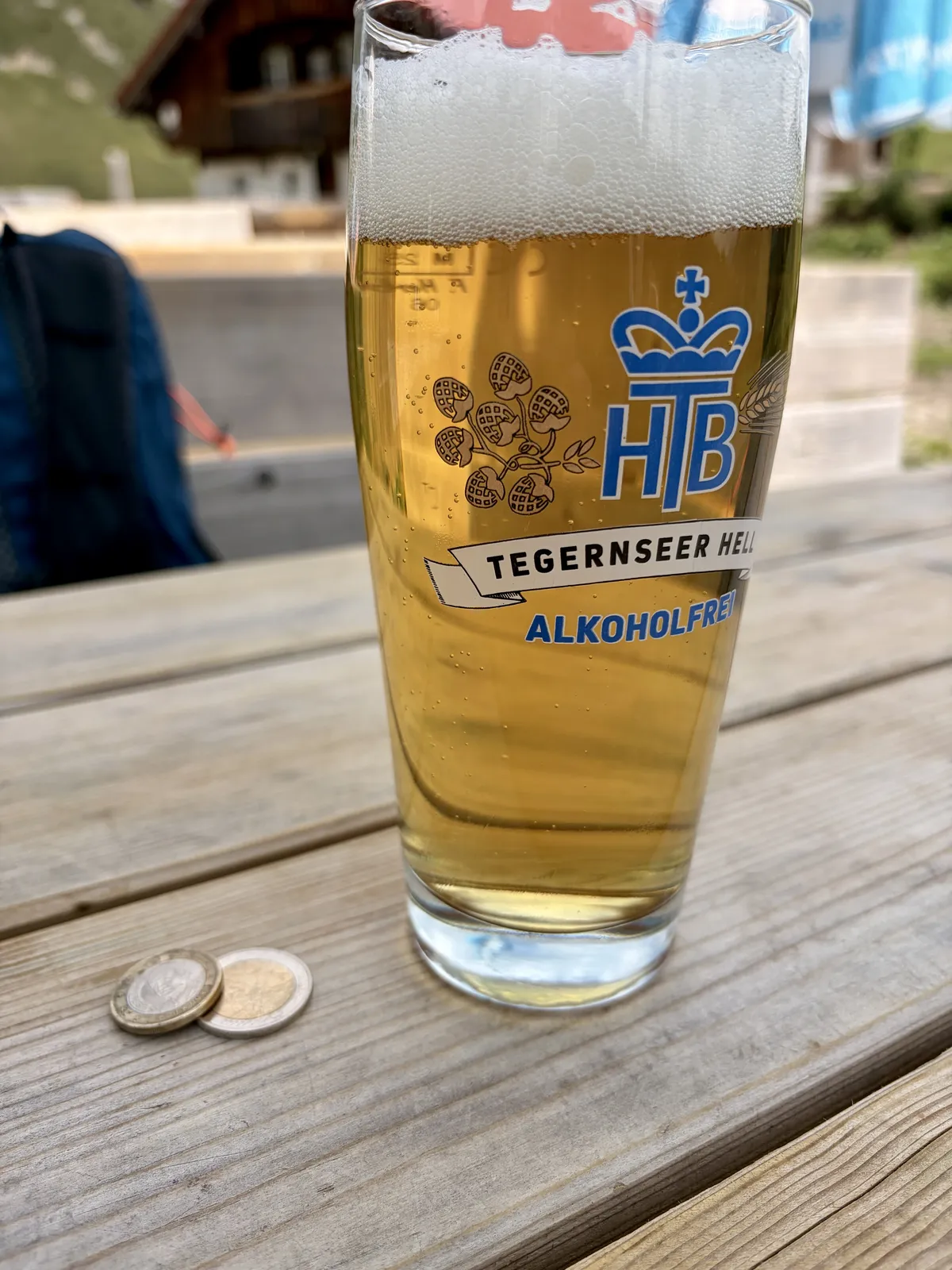 Ein Glas Tegernseer Helles Bier auf einem Holztisch, daneben liegen zwei Münzen. Im Hintergrund sind verschwommen eine Hütte und einige Fahnen zu sehen.