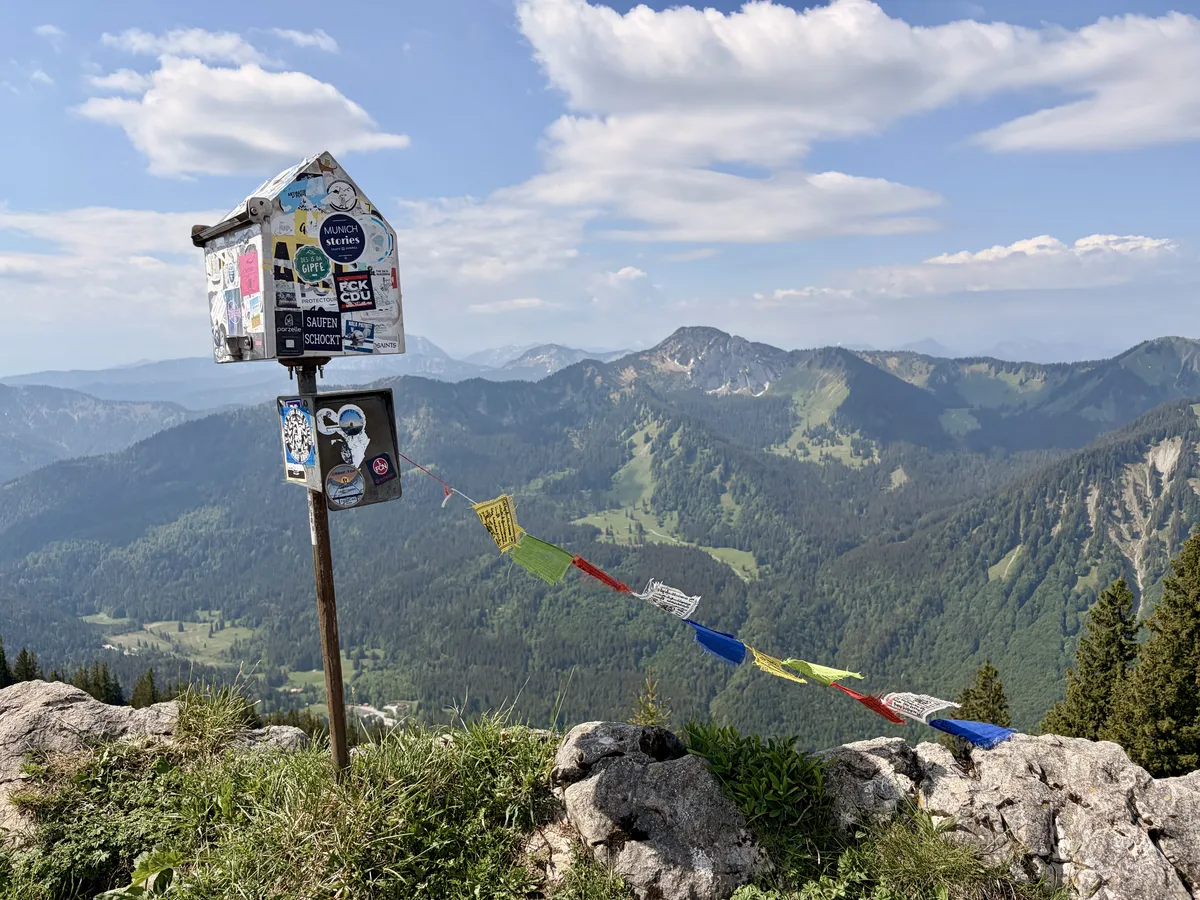 Ein kleiner Blechkasten mit Aufklebern steht auf einem felsigen Gipfel und verwahrt das Gipfelbuch, umgeben von einer beeindruckenden Berglandschaft. Bunte Gebetsfahnen wehen im Wind und verleihen der Szene einen spirituellen Charakter.
