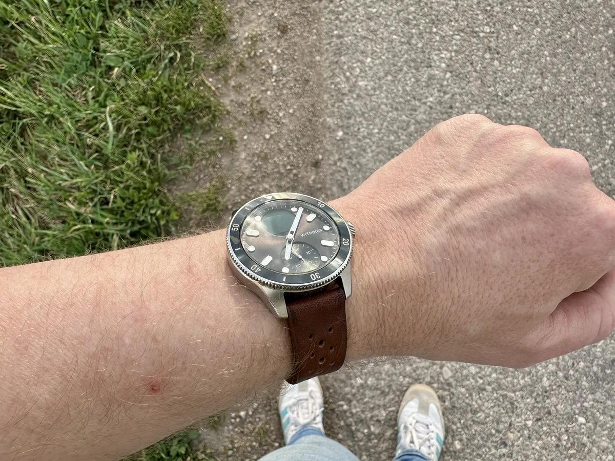 Ein Handgelenk mit einer Withings Nova und einem braunen Lederarmband. Im Hintergrund ist ein Kiesweg und grünes Gras sichtbar.