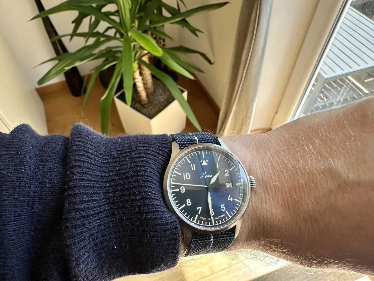 Ein Handgelenk mit einer Laco Stuttgart Pro mit blauem Zifferblatt und silbernem Gehäuse, das in einer Innenumgebung neben einer Pflanze abgebildet ist. Die Uhr zeigt die aktuelle Uhrzeit und hat ein Stoff-Armband.