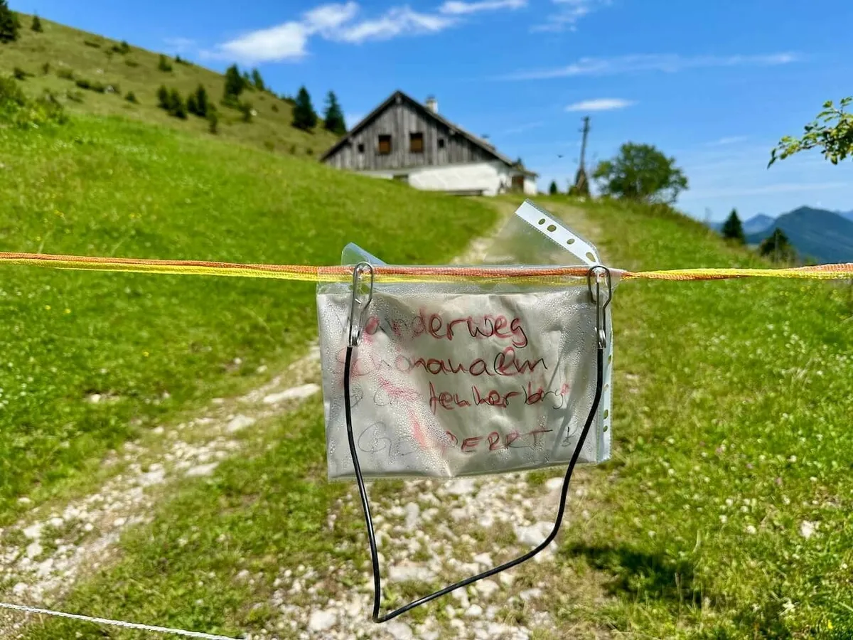 Durch ein Sperrband blockierter Wanderweg mit einem handgeschriebenen Hinweisschild ("Wanderweg gesperrt") vor einem Almhaus