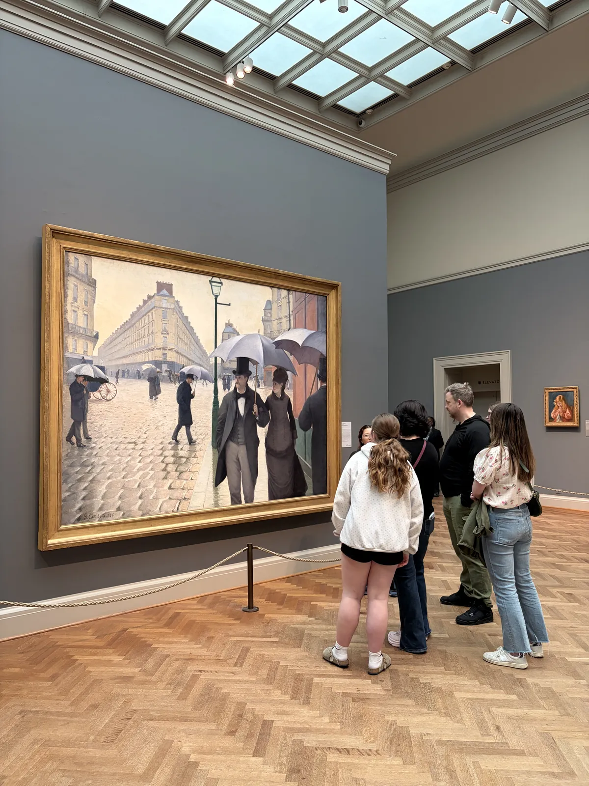 Eine Gruppe von Besuchern steht vor einem großen Gemälde, das eine belebte Straßenszene mit Menschen unter Regenschirmen zeigt. Der Raum ist hell erleuchtet und hat eine moderne Galerie-Atmosphäre.