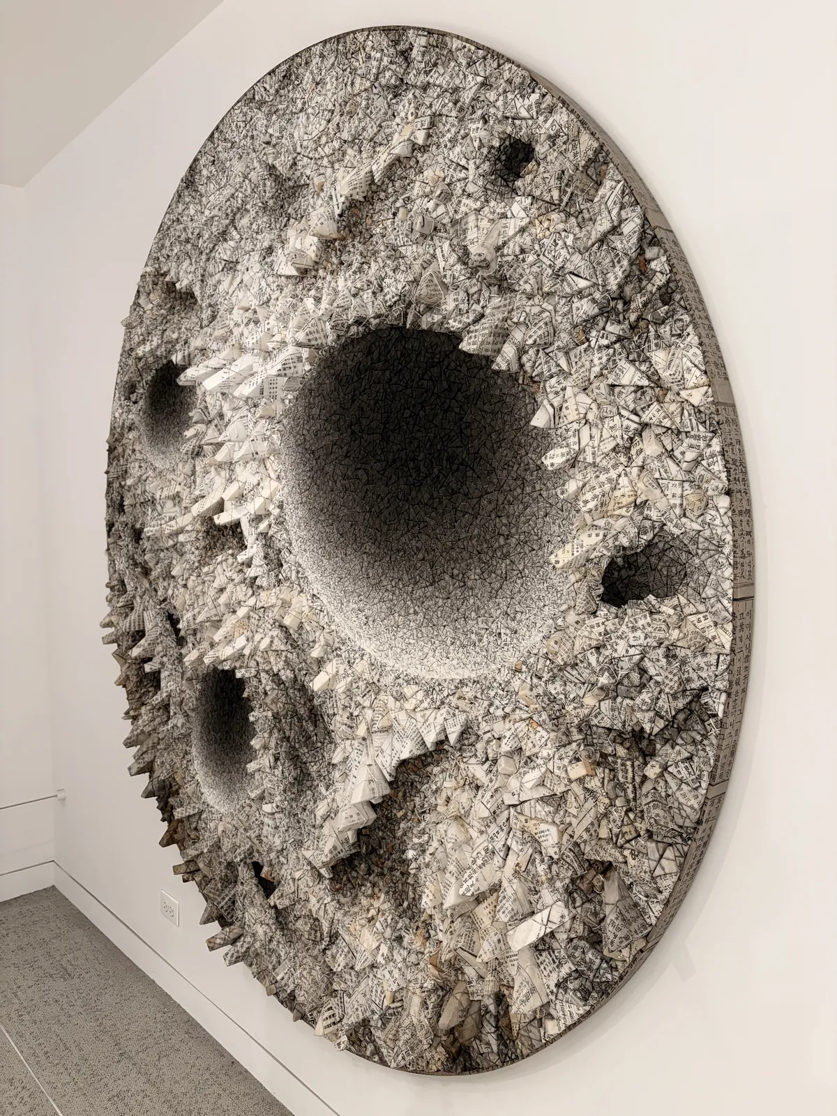 Eine große, runde Wandskulptur mit einer strukturierten Oberfläche aus grauen und weißen Materialien. Die Skulptur hat mehrere tiefe Vertiefungen und hervorstehende Formationen, die eine dreidimensionale Wirkung erzeugen.
