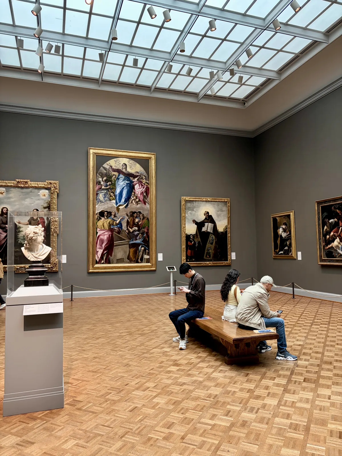 In einem Kunstmuseum sehen Besucher aus verschiedenen Blickwinkeln auf Gemälde, die an den Wänden hängen. Im Vordergrund sitzen zwei Personen auf einer Holzbank, während eine dritte Person auf ihr Handy schaut.