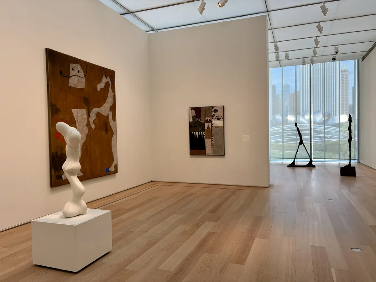 Im Inneren eines modernen Kunstmuseums beherbergt der Raum mehrere Kunstwerke, darunter eine abstrakte Skulptur auf einem weißen Sockel, eine gemalte Leinwand an der Wand und ein weiteres Gemälde daneben. Durch große Fenster ist die Skyline der Stadt sichtbar.