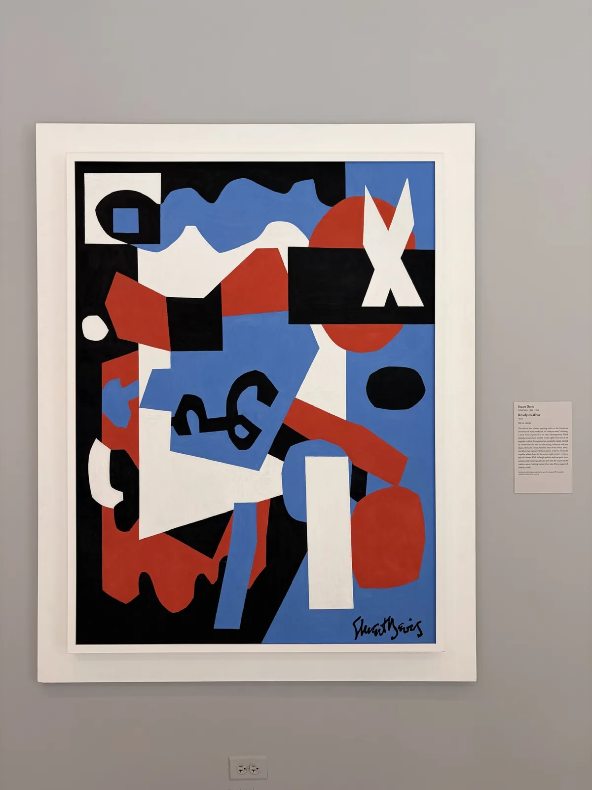 Ein abstraktes Kunstwerk mit kräftigen Farben, darunter Blau, Rot, Schwarz und Weiß. Es zeigt geometrische Formen und Zeichen, die dynamisch angeordnet sind, mit einer handschriftlichen Signatur am unteren Rand.