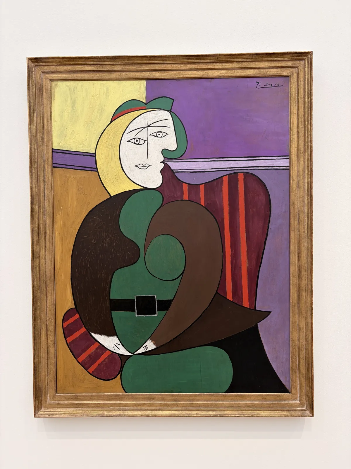 Ein farbenfrohes Gemälde von Pablo Picasso mit einer stilisierten Darstellung einer Frau. Die Frau hat blonde Haare, eine grüne Kleidung und sitzt auf einem Stuhl, umgeben von geometrischen Formen und kräftigen Farben.