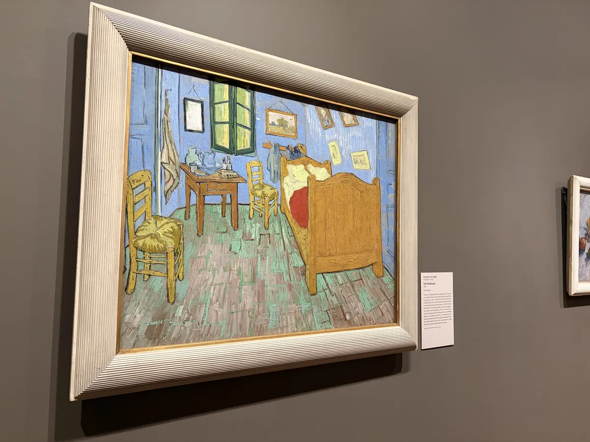 Ein Gemälde von Vincent van Gogh, das ein einfaches Schlafzimmer zeigt, mit einem Bett, einem Tisch und zwei Stühlen. Die Wände sind in Blau gehalten, und es gibt Fenster, die das Licht hereinlassen.