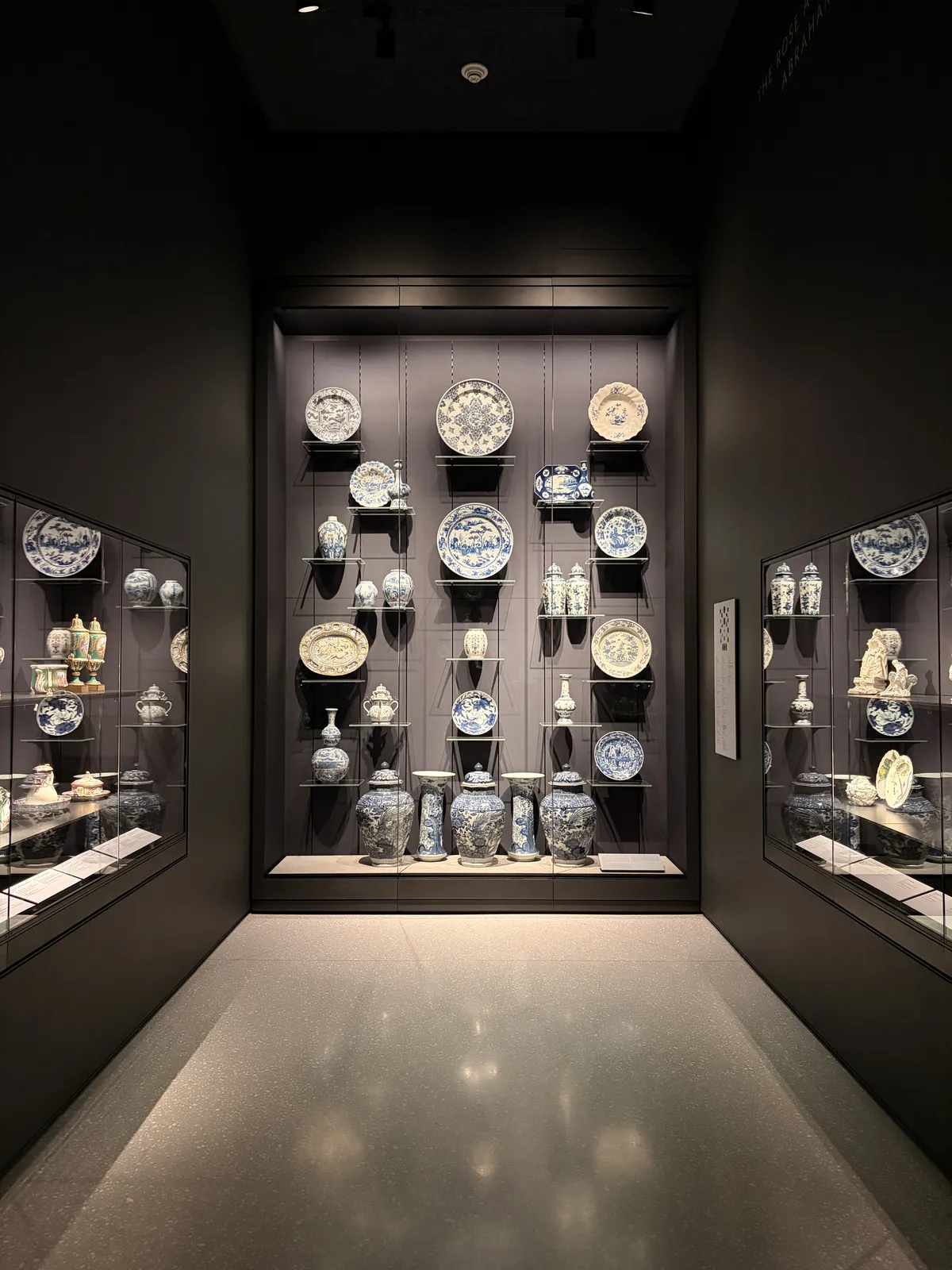 Eine dunkle Museumshalle mit zwei Vitrinen, die zahlreiche historische Porzellanobjekte zeigen. An der Wand hängen große Teller und Vasen mit blau-weißen Mustern, während die Vitrinen kleinere ornamente und Töpferwaren präsentieren.