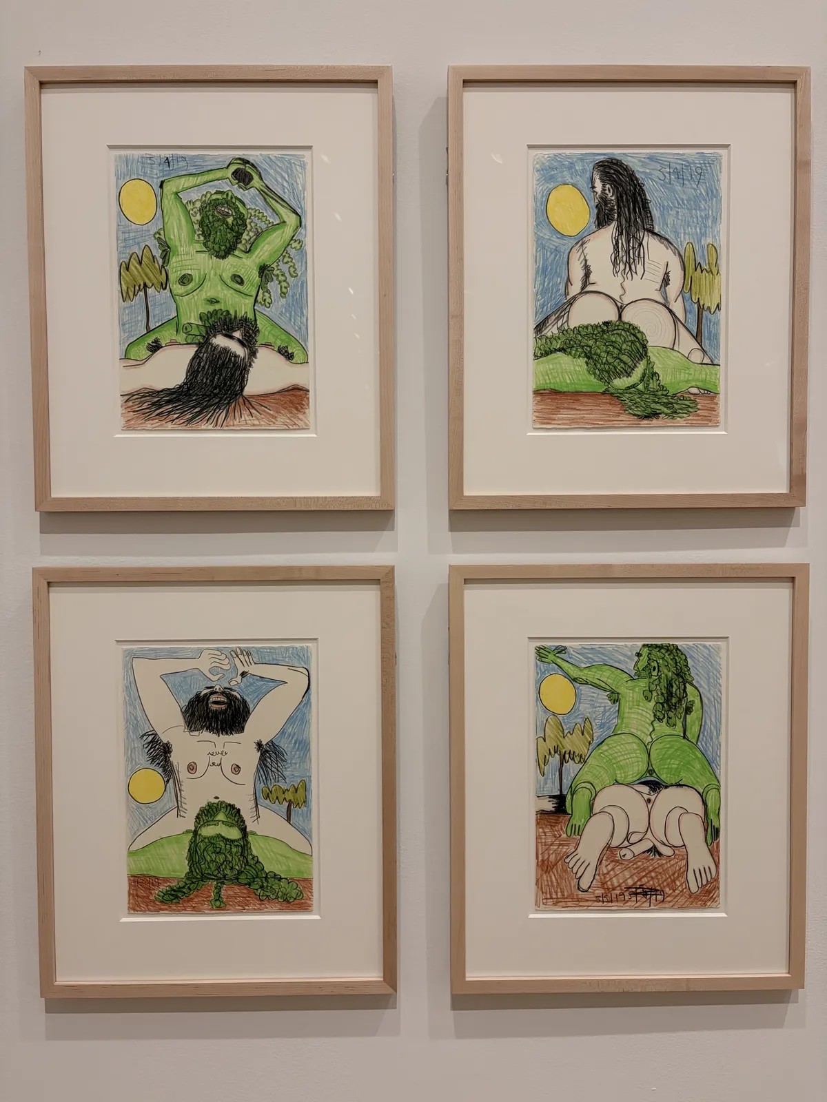 Vier gerahmte Illustrationen sind an einer Wand aufgehängt, die verschiedene Figuren in einem natürlichen Umfeld darstellen. Die Charaktere sind mit grünen und braunen Farben gestaltet und zeigen intime Posen zwischen Bäumen und einer gelben Sonne.