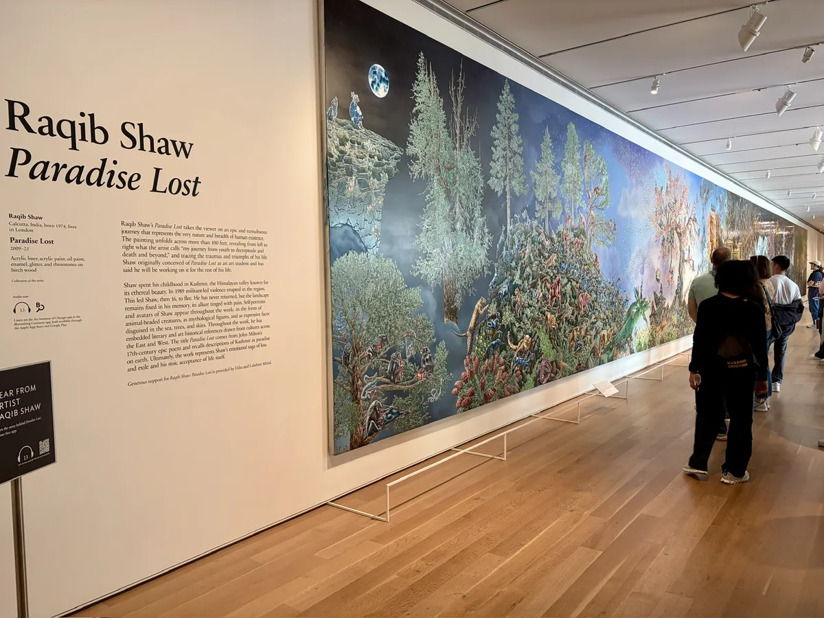 Eine lange Wandinstallation mit dem Titel "Paradise Lost" von Raqib Shaw, die eine farbenfrohe, detaillierte Landschaft darstellt, in der Menschen und Tiere in einer fantasievollen Umgebung interagieren. Besucher betrachten das Kunstwerk in einem modernen Ausstellungsraum mit hellem Holzfußboden.