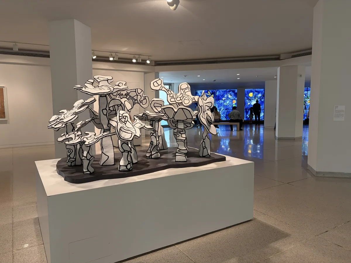 Eine skulpturale Installation mit stilisierten, schwarz-weißen Formen, die verschiedene Figuren darstellt. Im Hintergrund sind farbenfrohe Glasfenster und Besucher im Museum zu sehen.