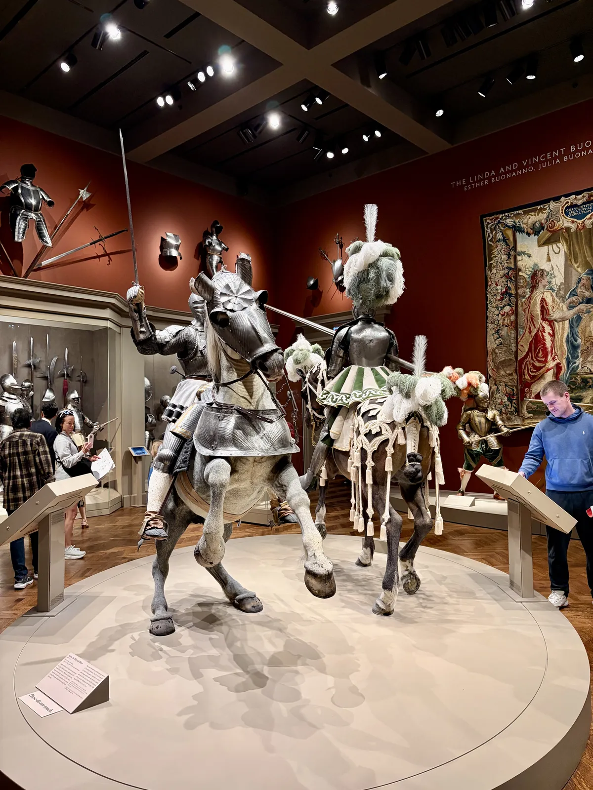 Zwei mit Rüstungen ausgestattete Ritter auf Pferden stehen in einem Museum. Der Hintergrund zeigt eine Wand mit weiteren ritterlichen Rüstungen und historischen Waffen, während Besucher das Exponat betrachten.