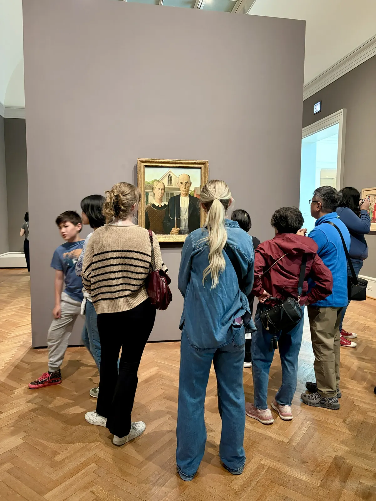 Eine Gruppe von Besuchern steht in einem Museum vor einem Gemälde, das ein Paar darstellt. Die Menschen sind unterschiedlich gekleidet und konzentrieren sich auf das Bild, während einige zu sprechen scheinen.