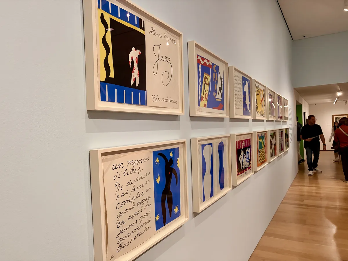 An Ausstellungswand zeigt eine Reihe von zwölf gerahmten Kunstwerken, die in bunten Farben und verschiedenen Stilen gestaltet sind. Einige Werke haben grafische Elemente und Texte, während Besucher die Kunstwerke betrachten.
