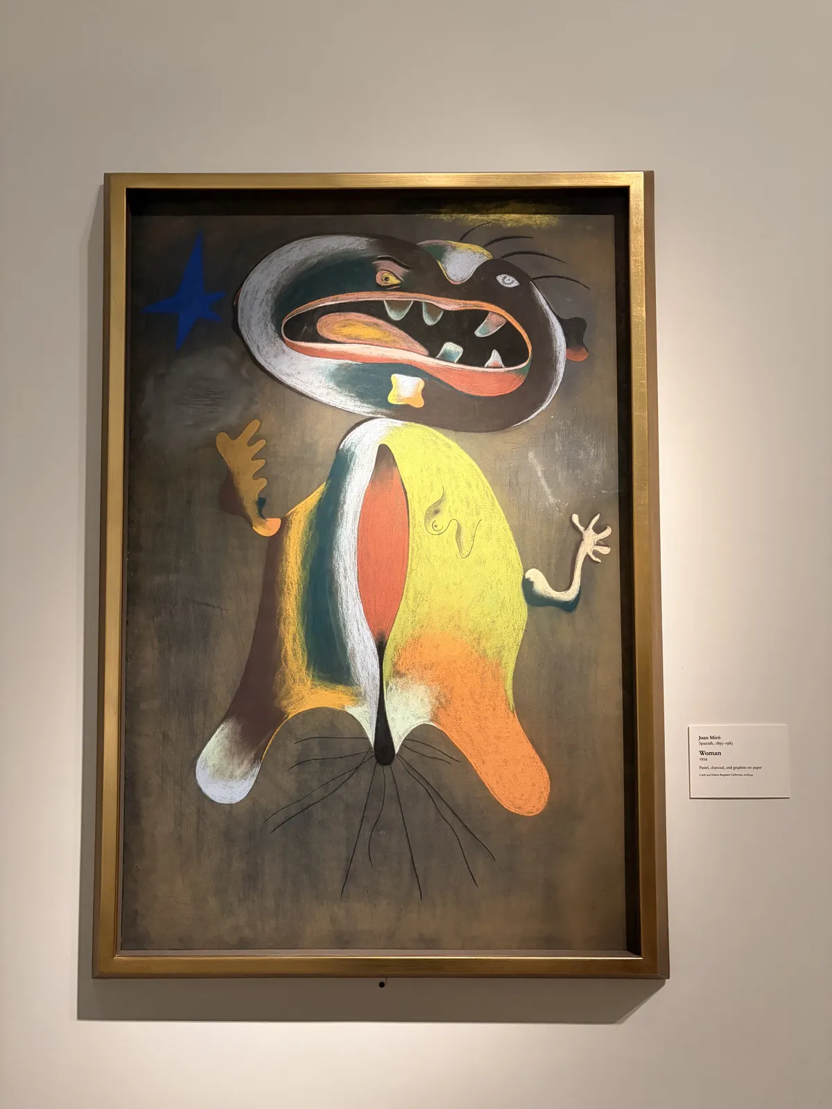 Ein farbenfrohes Gemälde zeigt eine cartoonartige Figur mit einem übertriebenen Gesichtsausdruck, die große Zähne und eine asymmetrische Körperform hat. Der Hintergrund ist dunkelfarbig, während die Figur leuchtende Farben in Grün, Orange und Gelb aufweist.